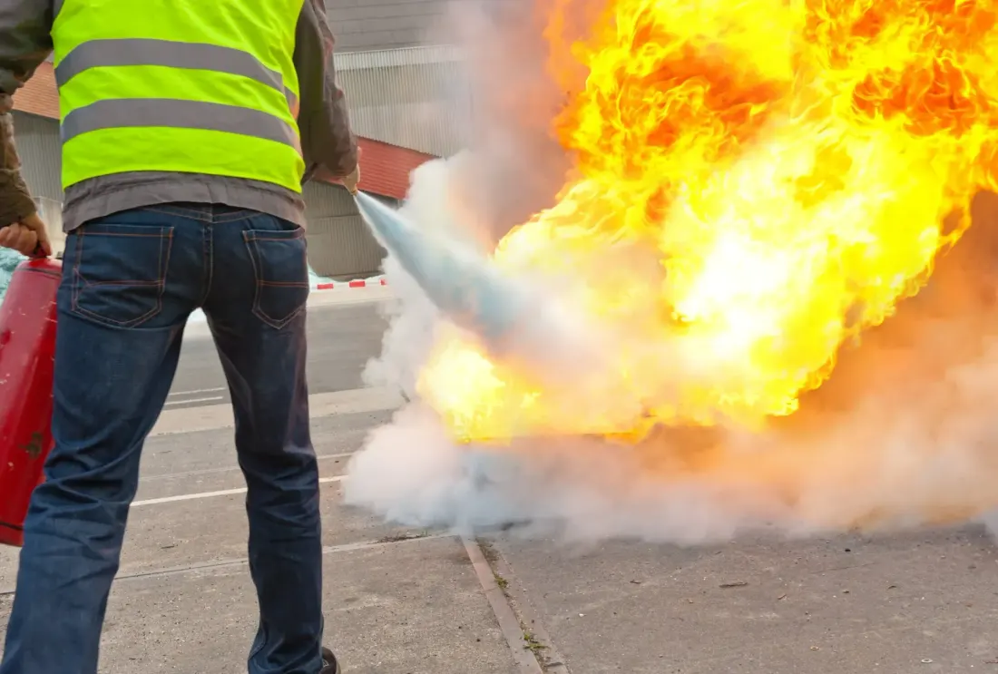 A person wearing a high-visibility vest uses a red fire extinguisher to suppress bright orange flames outdoors