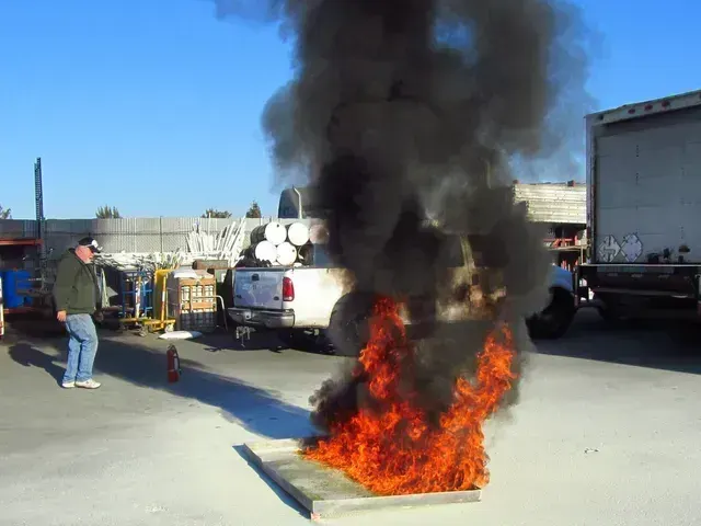 A person stands near a fire burning in a metal tray, producing a large cloud of thick black smoke