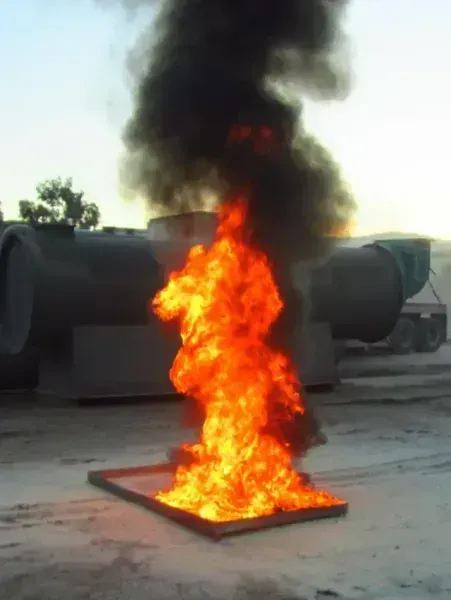A bright orange fire burns in a rectangular metal tray, producing thick black smoke outdoors near industrial tanks