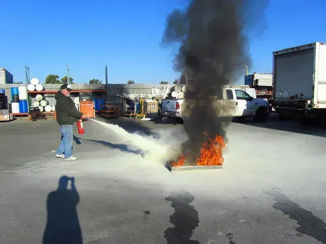 A person using a red fire extinguisher to put out a small, intense fire on a paved lot